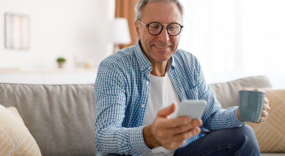 Image d’un homme souriant en chemise, assis sur un canapé avec une tasse de café et regardant son téléphone